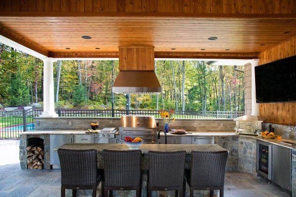 Outdoor kitchen with a wooden ceiling, grill, stone countertops, woven chairs, and a forest view in the outdoor background