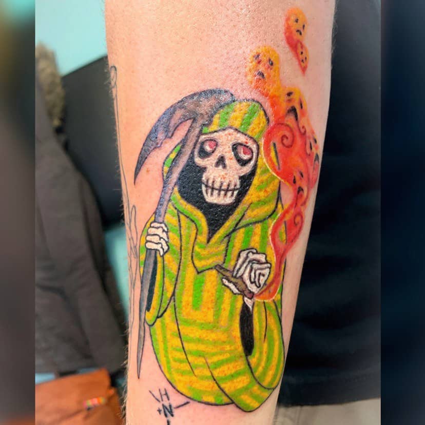 A colorful tattoo of a grinning Grim Reaper in a green-striped robe, holding a scythe, with fiery ghostly faces swirling around.