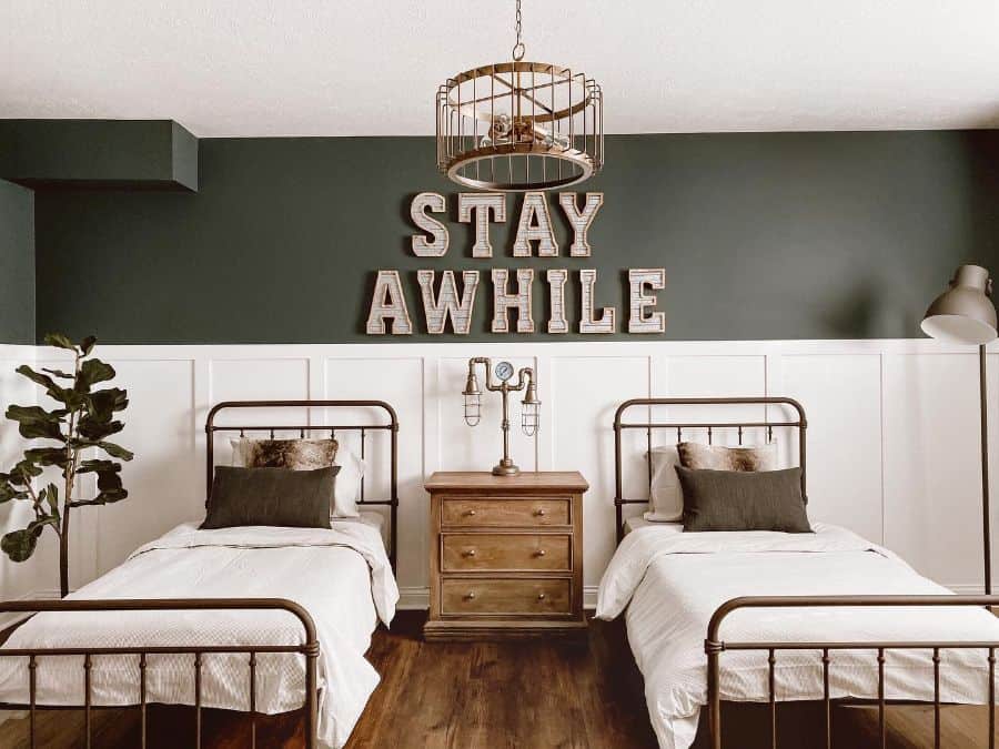 Rustic twin bed guest room with dark green accent wall, wooden decor, and "Stay Awhile" sign.