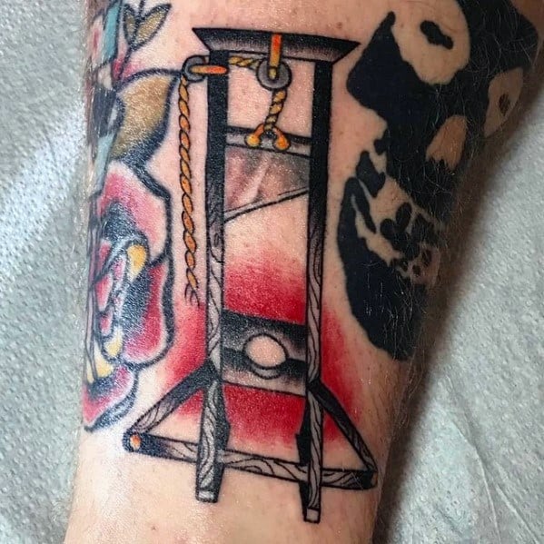 Guillotine tattoo with bold black outlines, red shading in the background, and surrounding elements like a rose and skull design.