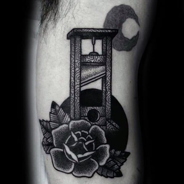 Blackwork guillotine tattoo with ornate patterns, a dark circular backdrop, and a detailed rose blooming at the base.