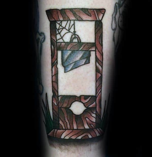 Colorful guillotine tattoo with wood grain pattern, a blue blade, cobweb detail, and grass accents at the base.