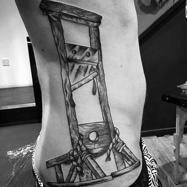 Large black and grey guillotine tattoo along the ribs with wood texture, dripping details, and thick rope elements tied at the base.