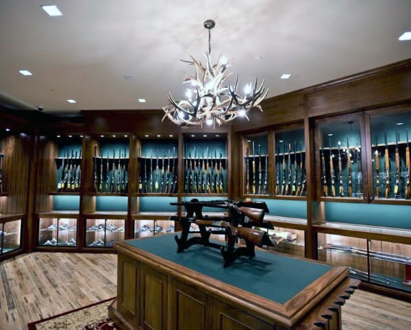 Elegant gun room featuring custom wooden cabinetry, glass displays, and a central island for showcasing firearms, illuminated by an antler chandelier.