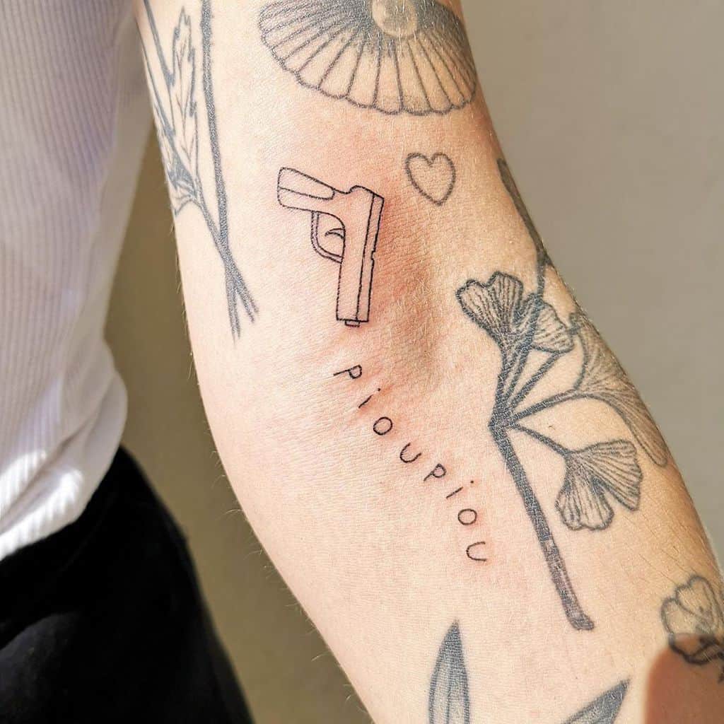 A minimalist black ink tattoo of a small gun with the word 'pioupiou' resembling a playful sound effect, placed on the forearm.