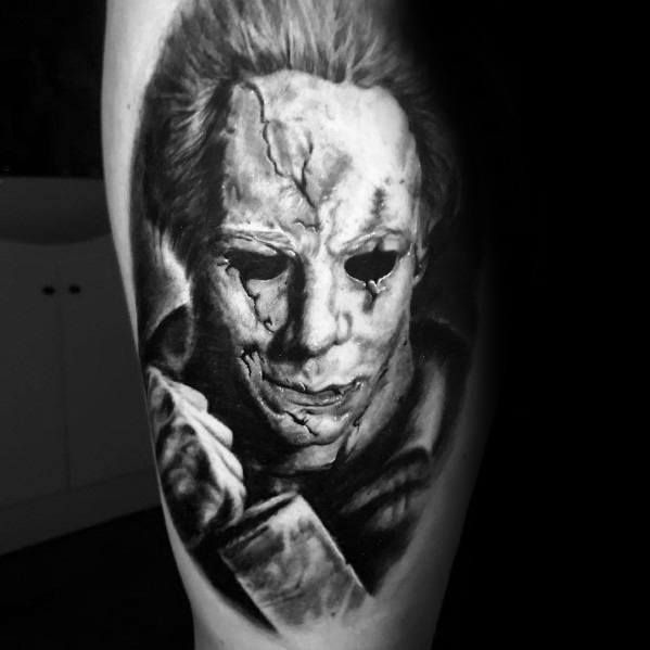Black and grey Michael Myers tattoo with a cracked mask and menacing stare, holding a knife for a terrifying horror effect.