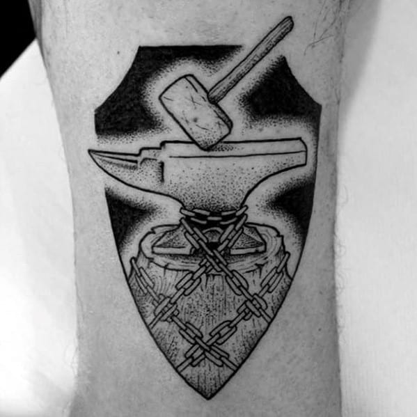 Black and white anvil tattoo on a stump wrapped in chains, with a hammer above, all framed within a shield-shaped background.