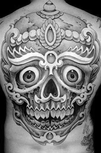 Black and grey Tibetan skull tattoo with wide eyes, ornate crown, and bold shading, covering the upper to mid back.