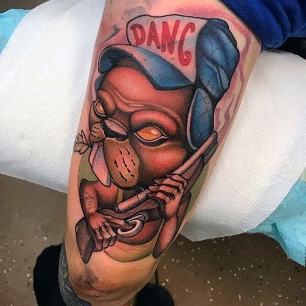 Cartoon-style beaver tattoo wearing a “DANG” cap, holding a rifle with a tough expression and chewing on a twig.