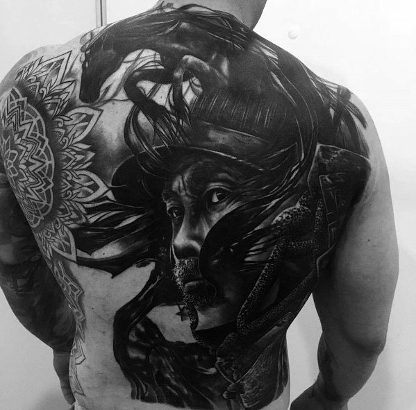 Large full-back tattoo in black and grey featuring a realistic warrior’s face, flowing hair, a snake, and intricate mandala patterns.