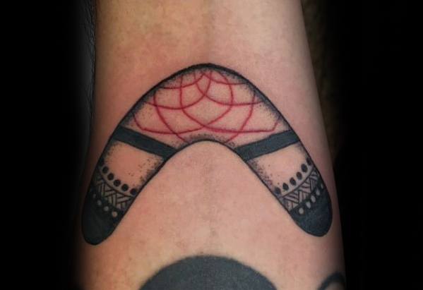 Black boomerang tattoo with tribal-style ends and a red dreamcatcher web design in the center, placed on the inner forearm.