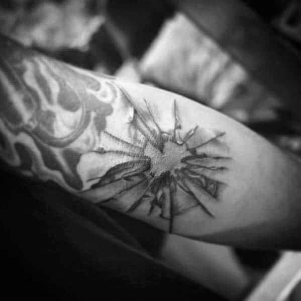 Minimalist black and grey tattoo of a realistic bullet hole-style shattered glass effect on the inner forearm.