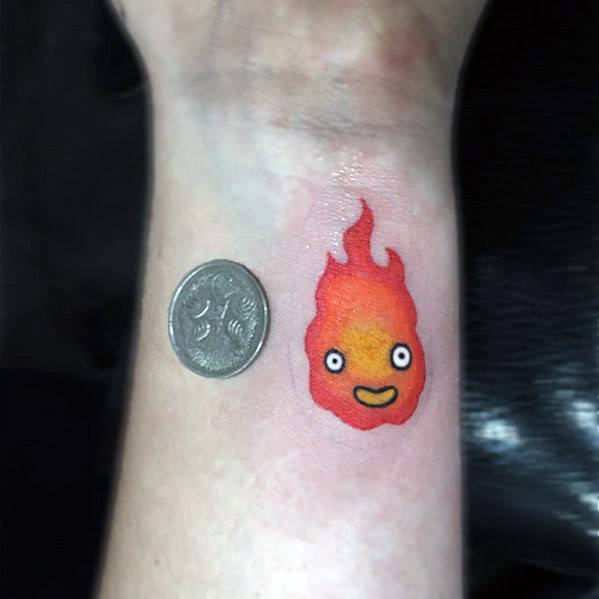 Tiny Calcifer wrist tattoo with a warm red-orange flame, smiling face, and a coin beside it for size comparison.