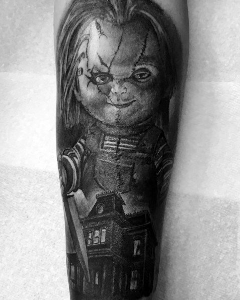 Black and grey Chucky tattoo featuring his sinister grin, stitched face, and a knife, with a haunted house below for a chilling horror design.