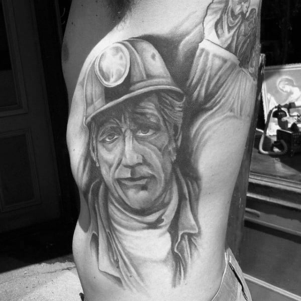 Realistic black and grey tattoo of a coal miner with a helmet and headlamp, featuring a tired but determined expression.