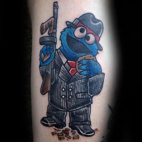 Gangster-style Cookie Monster tattoo in a pinstripe suit, holding a cookie and tommy gun with bullet casings scattered below.