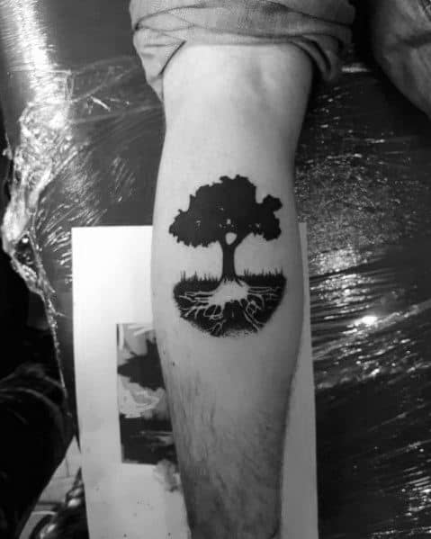 Bold black ink tattoo of a tree with deep roots inside a circular design, symbolizing strength, growth, and connection to nature.