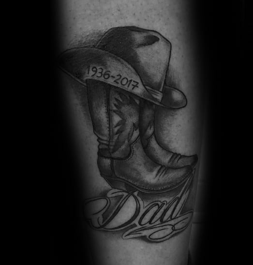Black and grey tattoo of cowboy boots with a hat inscribed "1936-2017" and "Dad," serving as a heartfelt tribute and memorial piece.