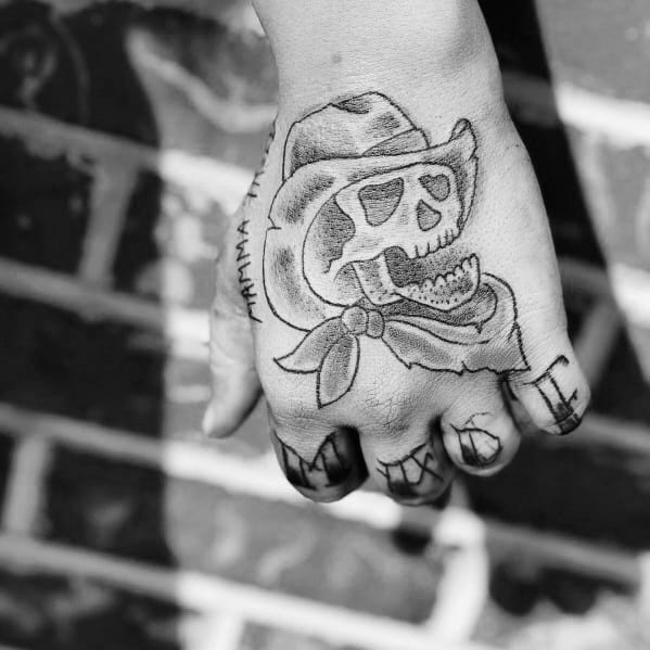 Black ink tattoo of a grinning cowboy skull wearing a hat and bandana, placed on the hand with bold linework and a rugged Western feel.