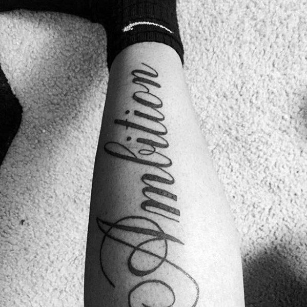 A tattoo of the word "Ambition" in elegant cursive on a leg resting on carpet, with a sock partially visible