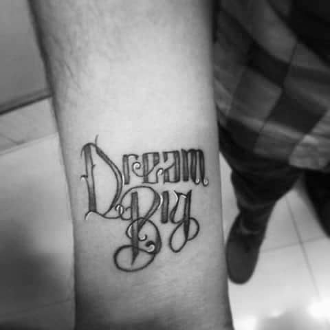 Forearm tattoo in decorative blackletter script that reads “Dream Big,” combining bold uppercase and elegant cursive elements.
