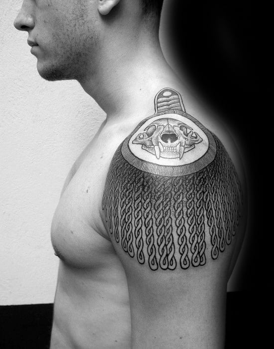 Epaulette tattoo with a bold saber-toothed skull centerpiece and long twisted tassels flowing over the shoulder.