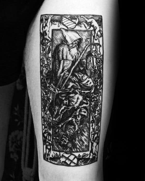 Tattoo resembling an intricate tarot card, featuring a hooded figure with a staff, surrounded by symbolic animals and ornate border details.