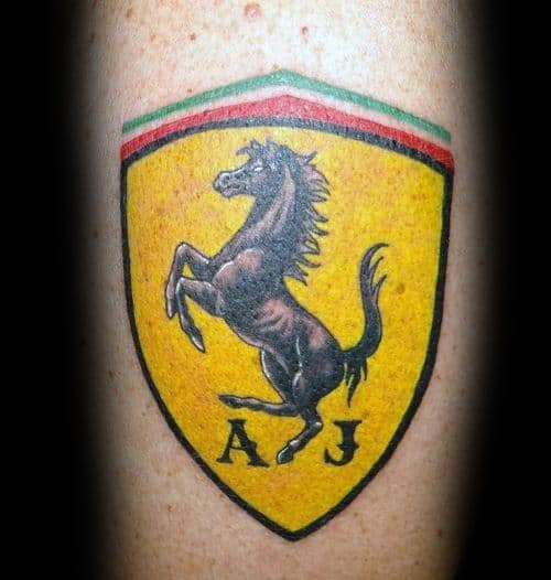 Classic Ferrari shield tattoo featuring a black prancing horse on vibrant yellow, personalized with initials "A J," inked on man's arm.