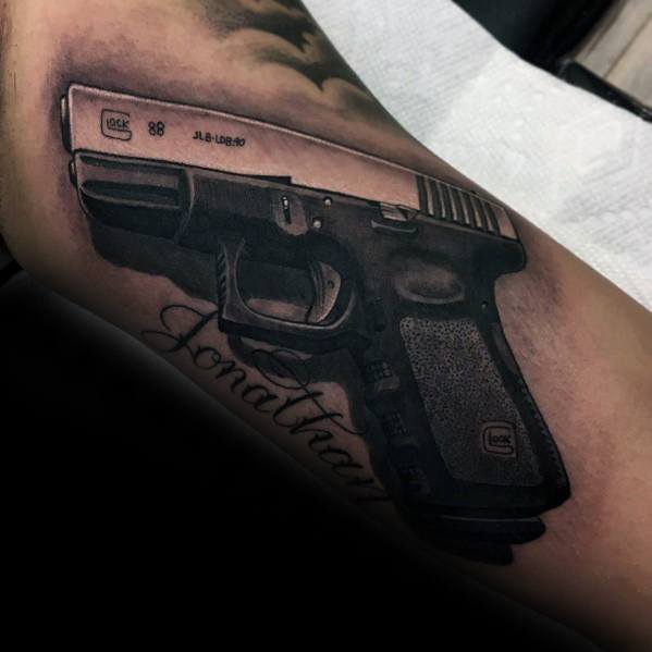 Realistic black and grey tattoo of a Glock 98 on the arm, featuring detailed shading and the name "Jonathan" elegantly scripted below.