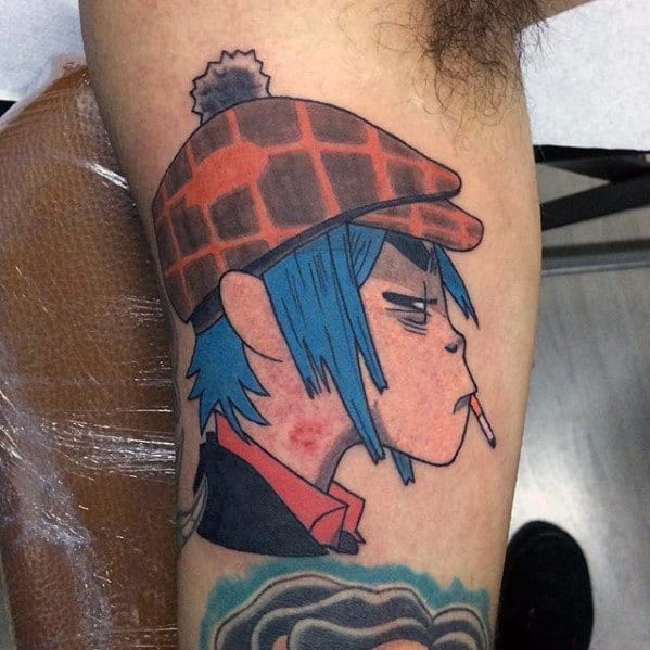 Color tattoo of Murdoc from Gorillaz in profile, wearing a red plaid hat, blue hair, cigarette in mouth, and visible lipstick marks on neck.