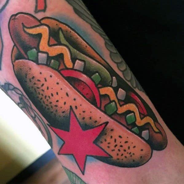Hot dog tattoo featuring mustard, onions, and tomatoes inside a bun, with a red star symbol on the side, on the forearm.