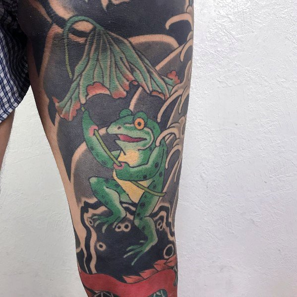 Japanese-style tattoo on the thigh featuring a green frog holding a leaf umbrella, surrounded by swirling water and traditional elements.