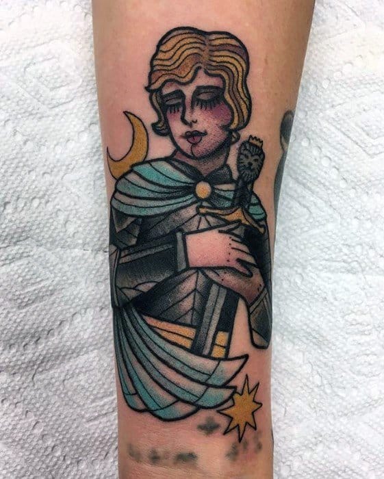 Traditional style Joan of Arc tattoo on the forearm with bold lines, muted tones, a sword, crescent moon, and star details.