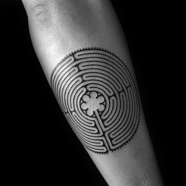 Circular labyrinth tattoo on a man's forearm, featuring a detailed maze design with a four-leaf clover at the center and a bold black outline.