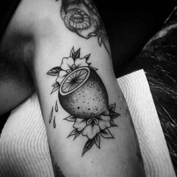 Black and grey tattoo of a halved lemon with juice droplets, framed by detailed flowers and leaves in a classic style.