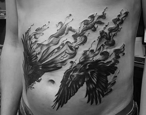 Dynamic black and grey tattoo of Odin’s ravens in flight, transforming into smoke across the lower ribs and stomach.
