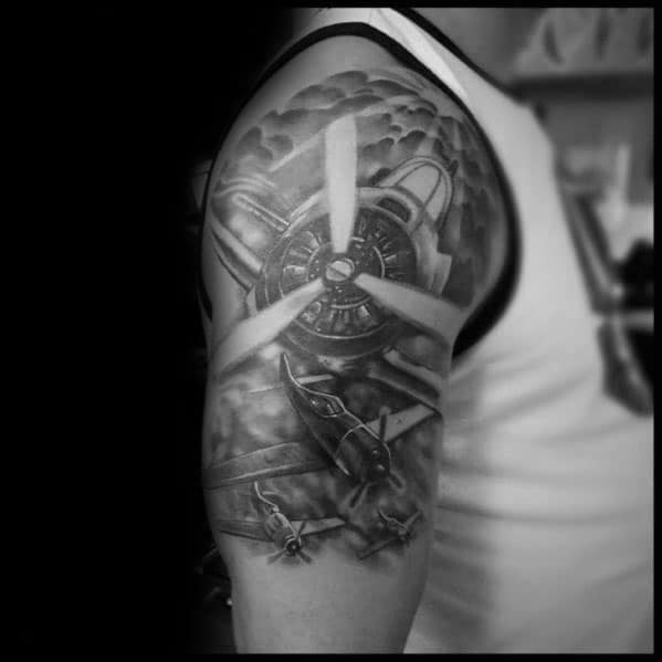 Realistic black and grey aviation-themed sleeve tattoo featuring a large propeller engine, fighter planes in flight, and dramatic cloud shading.