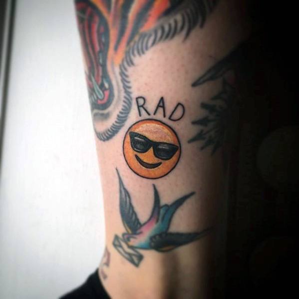 Tattoo of a smiling sunglasses emoji with the word "RAD" above it, surrounded by other colorful tattoos