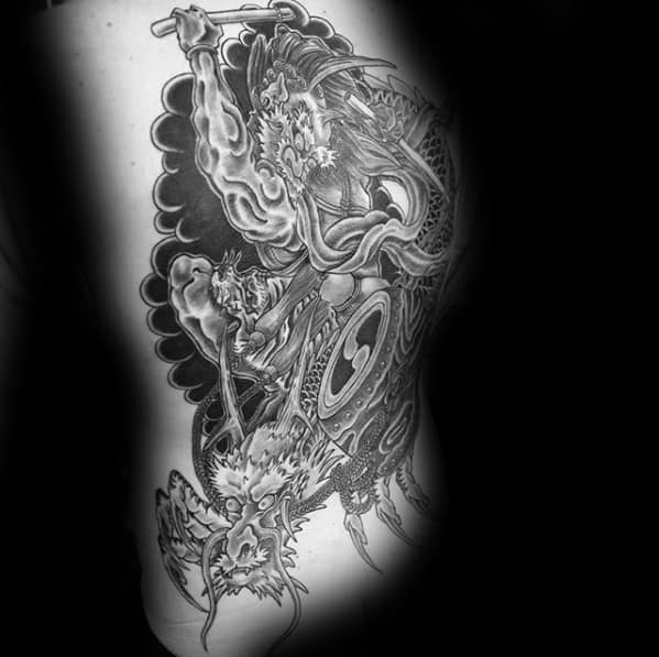 Black and grey Raijin side torso tattoo with dynamic thunder god, swirling clouds, drums, and a fierce dragon in traditional style.