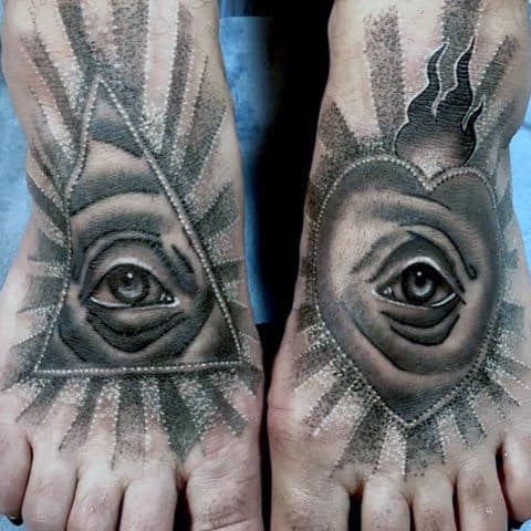 Two feet with detailed tattoos of eyes, one in a triangle and the other in a heart, both surrounded by rays