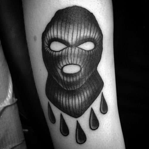 Tattoo of a dark ski mask with six large black teardrops falling beneath it, placed vertically on the arm.