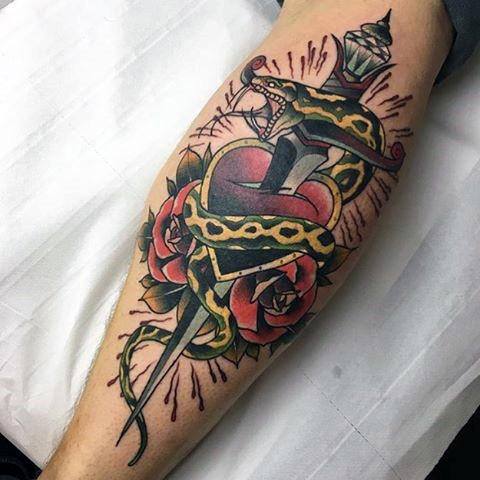 Traditional snake and dagger tattoo on leg, featuring a heart, roses, and bold black, red, and yellow ink with intricate shading.