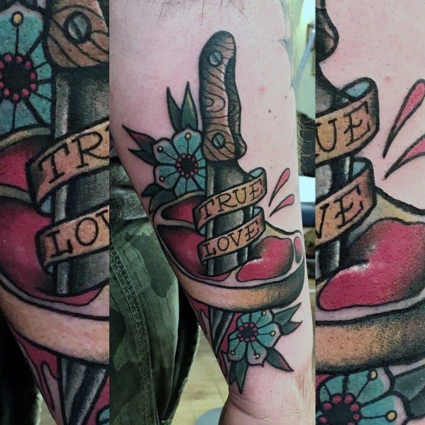 Tattoo of a steak pierced by a dagger, wrapped in a banner reading true love, with floral accents and bold shading for a classic tattoo style