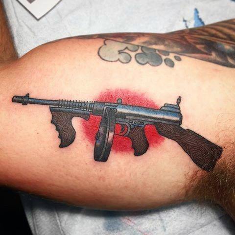 Realistic Tommy gun tattoo with bold black and grey shading, accented by a red background for a striking contrast on the upper arm.