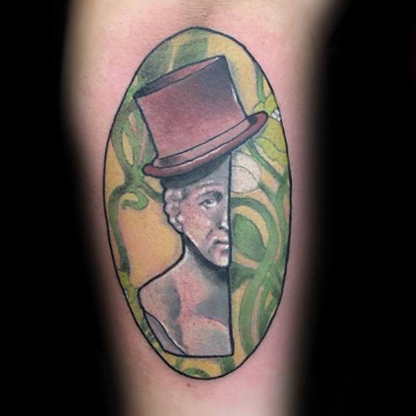 Abstract Willy Wonka tattoo with a split-face design, red top hat, and swirling green and yellow background inside an oval frame.