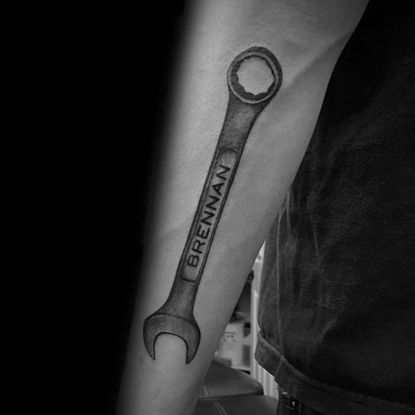 Realistic black and grey tattoo of a wrench on the forearm with the name BRENNAN engraved down the handle.