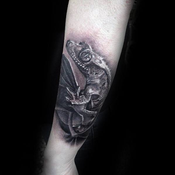 Hyper-realistic gecko tattoo on forearm with fine shading and texture, clinging to a leaf with a dark, moody background.