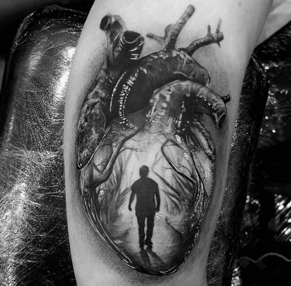 Realistic black and grey tattoo of an anatomical heart with a silhouette of a person walking inside, symbolizing emotion or inner journey.