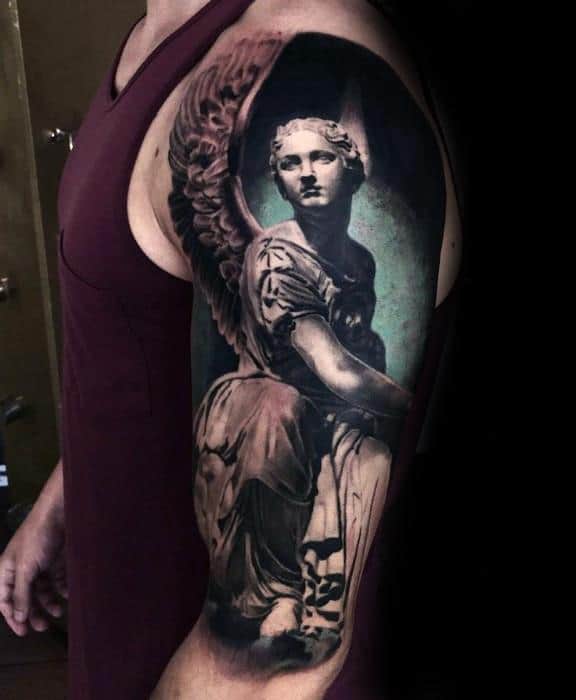 A detailed black and grey tattoo on the upper arm featuring an angel in a seated position, with large wings and a serene expression, set against a softly shaded background.