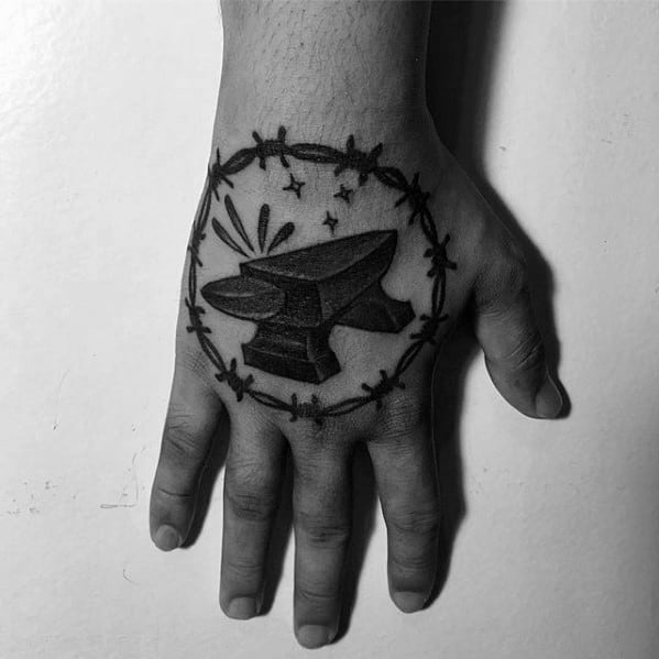 Bold black anvil tattoo on the hand, framed with barbed wire and accented by stars and spark lines for a striking, symbolic look.